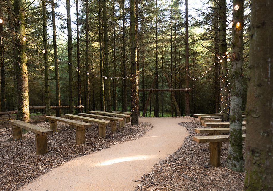 Wooden benches facing a wooden arch at the end of a path with trees on either side of the benches and fairylights above