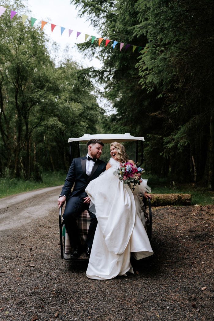 a bride and groom sitting on the back of a white golf buggy in a woodland setting