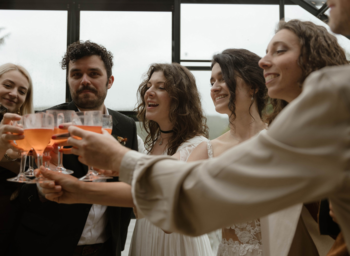 a group of people at a wedding, including two brides, standing in a circle and reaching in to toast wine glasses filled with an orange coloured drink