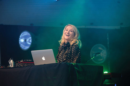 Blonde smiling DJ with apple macbook against a black backdrop