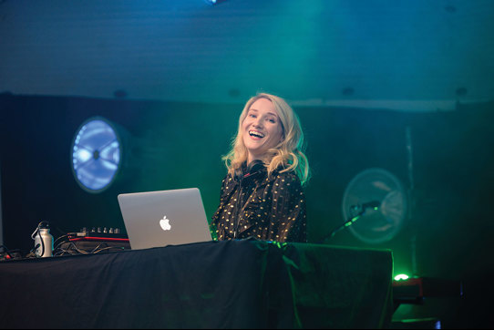 Blonde smiling DJ with apple macbook against a black backdrop