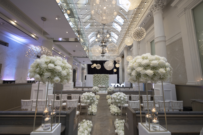 A long room with skylight windows and white pillars along each wall set up for a wedding ceremony with white floral arrangements