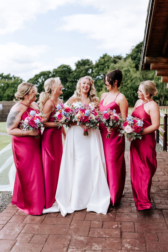 a bride with four bridesmaids wearing long hot pink slip dresses. They are all holding colourful bouquets of flowers and there are trees in the background