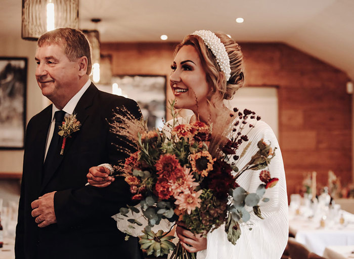 a happy bride holding a bouquet of dried flowers walking on the arm of an older man