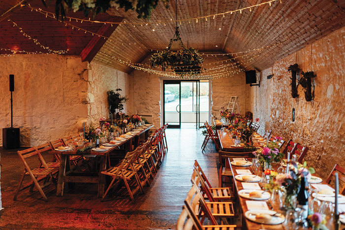 a rustic barn set with long wooden tables and chairs set for a wedding dinner.