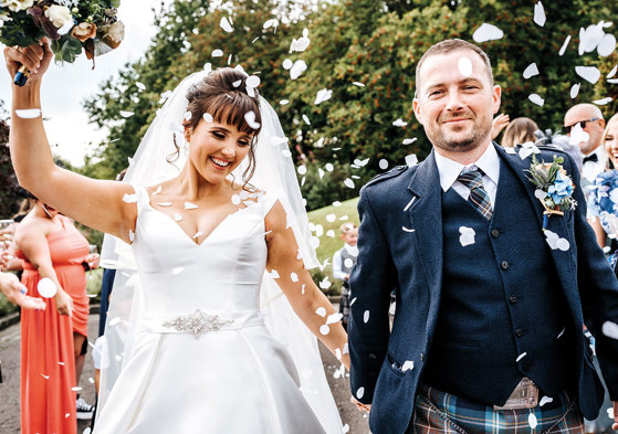 bride and groom walk towards camera smiling while their guests throw white coloured confetti on them