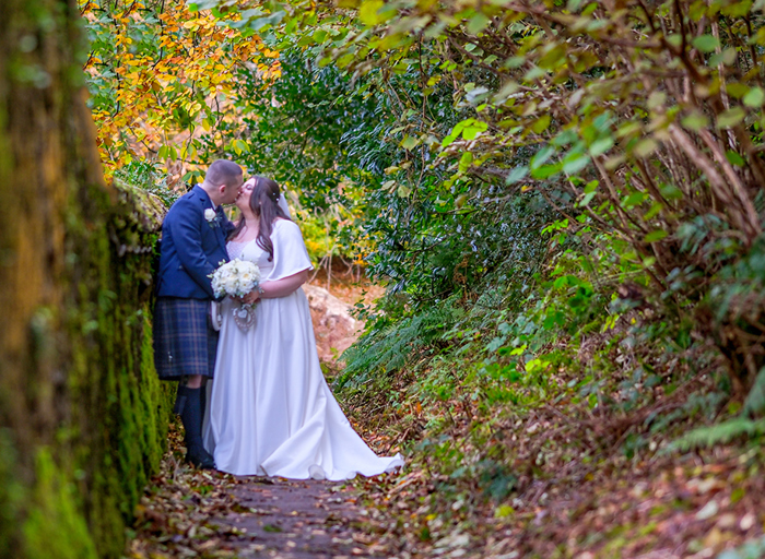 a bride and groom stand on a path kissing in a woodland area