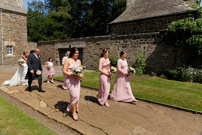 A bridal party walk along a stone path across the camera 