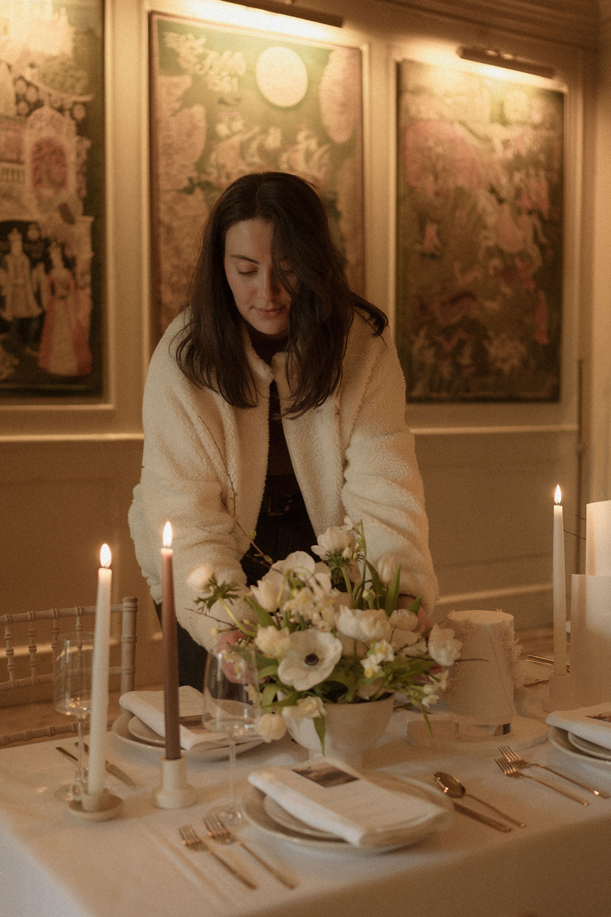 brunette woman stands in dimmed light, arranging a vase of neutral flowers on a tablecloth set dining table, complete with candlesticks, wine glasses, and multiple place settings