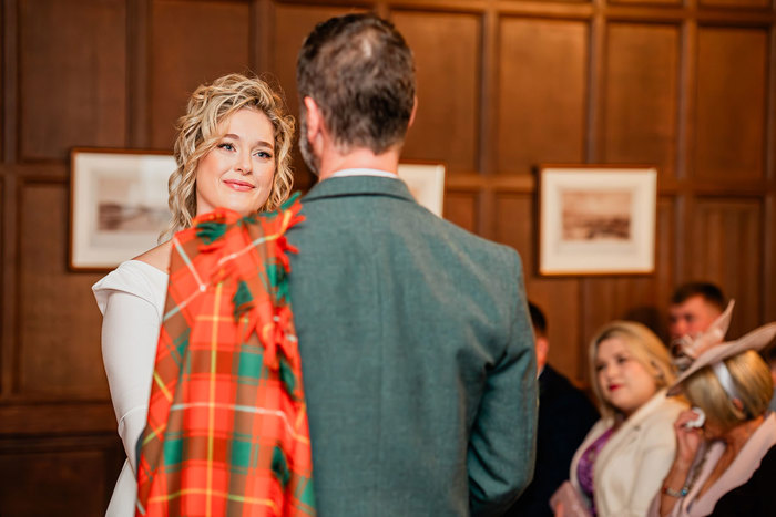 bride in white wedding dress looks lovingly at groom in red and green tartan kilt outfit