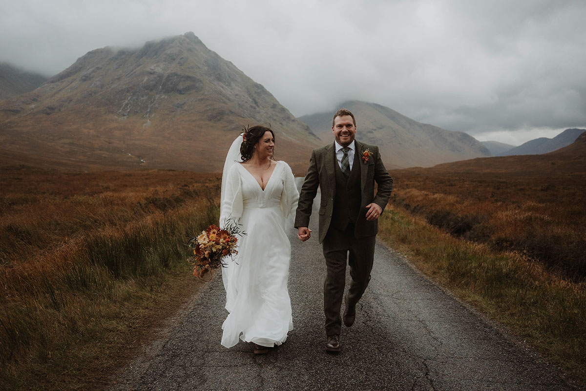a bride and groom walking along a narrow road in the shadow of Buachaille Etive Mòr