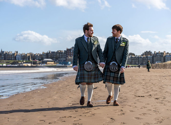 two grooms wearing green tweed kilt outfits walking hand and hand along a sandy beach with St Andrews skyline in the background