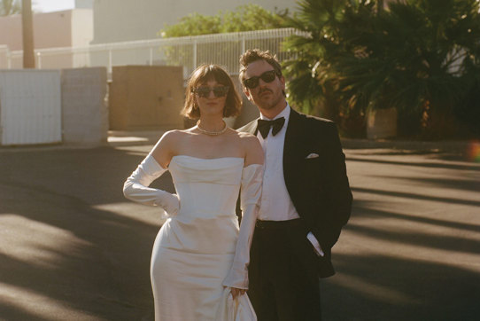 A couple dressed in wedding attire stands together outdoors, with palm trees and a building in the background