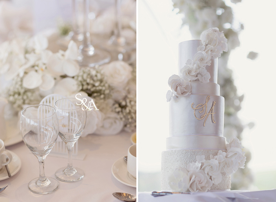 a wedding table setting with wine glasses on left. A white lustre wedding cake with white flower decor and gold letter monogram on right