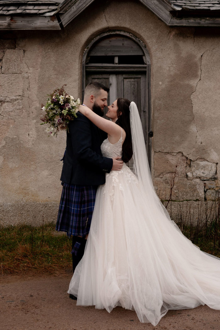 a bride and groom kissing in front of a worn wooden door. The building behind them has cracked walls and worn woodwork