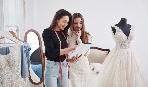 dark brunette woman with pink measuring tape hanging around her neck holds notebook and chats to another brunette woman in a wedding dress, pointing at the notebook as they stand in a changing room with other wedding dresses on display