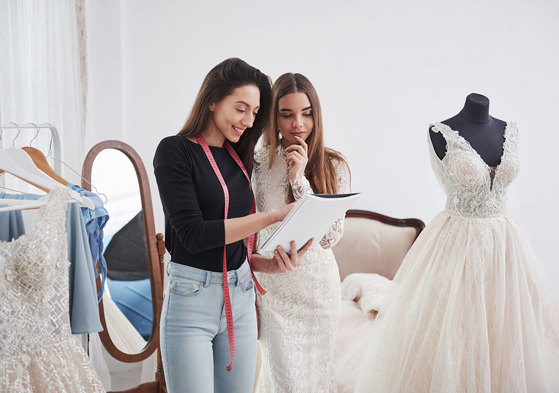 dark brunette woman with pink measuring tape hanging around her neck holds notebook and chats to another brunette woman in a wedding dress, pointing at the notebook as they stand in a changing room with other wedding dresses on display