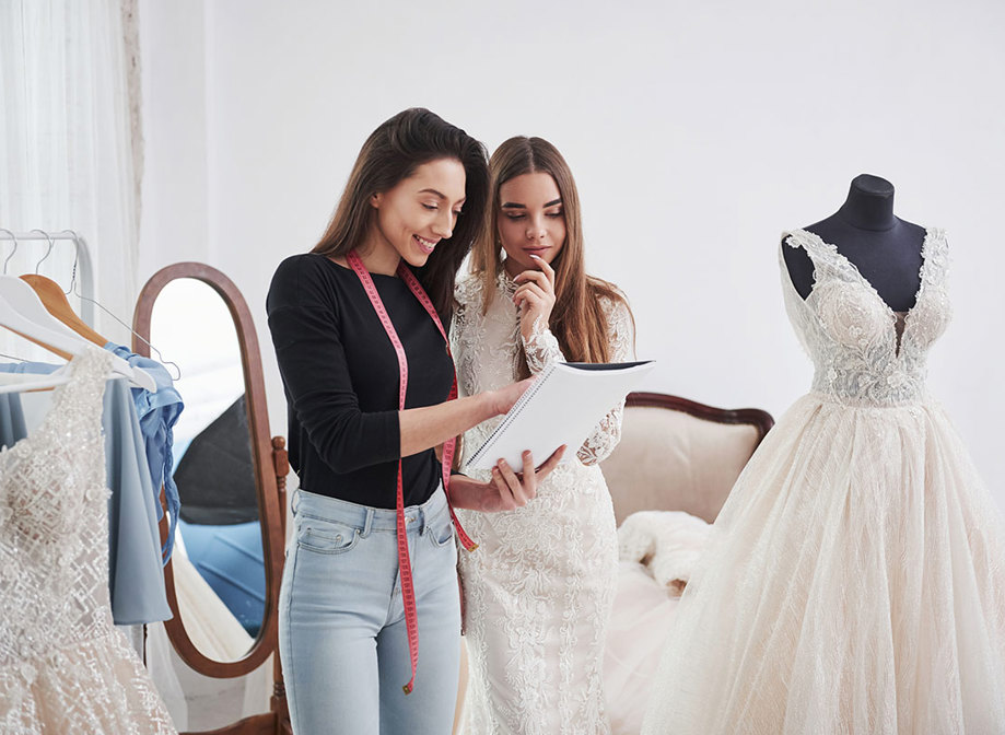 dark brunette woman with pink measuring tape hanging around her neck holds notebook and chats to another brunette woman in a wedding dress, pointing at the notebook as they stand in a changing room with other wedding dresses on display