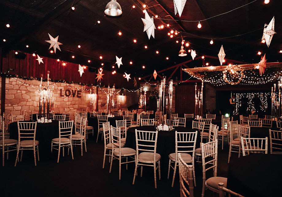 A room with brick walls and round tables with black tablecloths with white chiavari chairs around the tables. Fairy lights and white stars hang from the ceiling