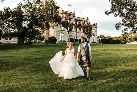 A bride and groom walk holding hands across grass away from a country house