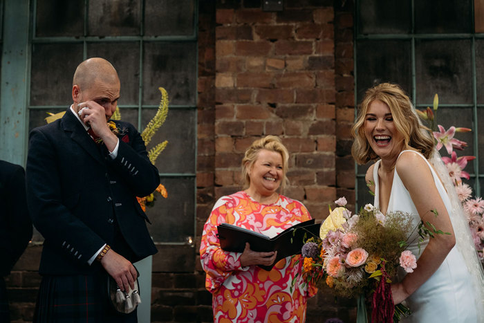 a bride laughing and groom wiping a tear away during a wedding ceremony officiated by Humanist Society Scotland. The celebrant is wearing a bright orange and pink patterned dress