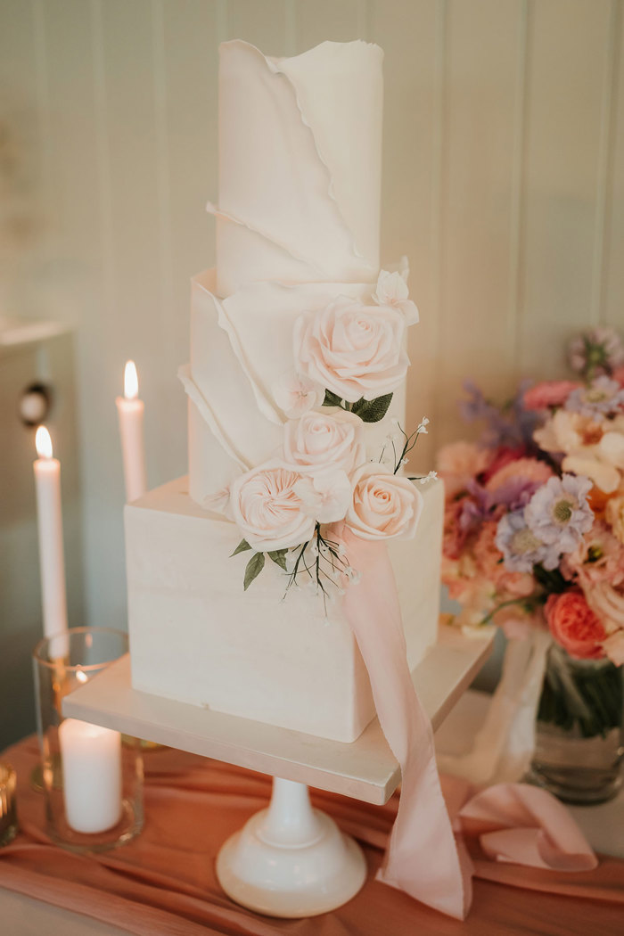 A three tier wedding cake with a square base and two round tiers on top and pale pink roses decorating one edge 