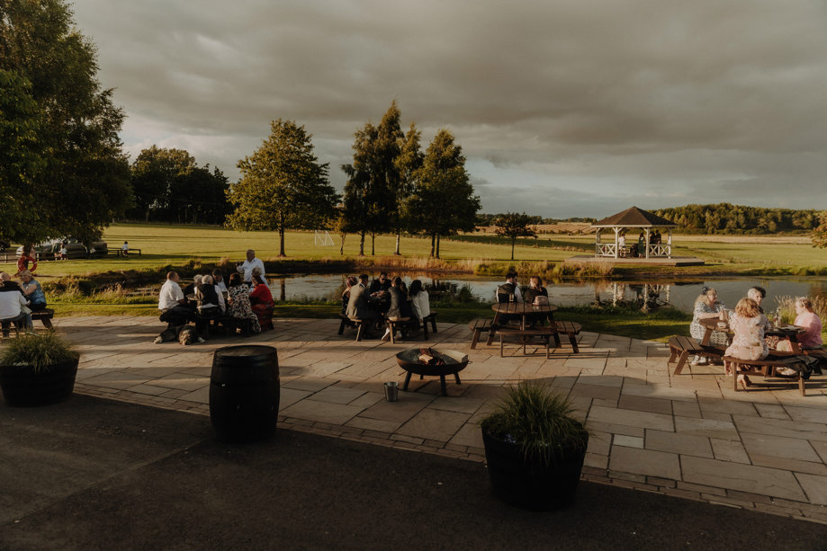 guests sit outside on communal benches talking to one another during wedding at bachilton barn as the sun goes down