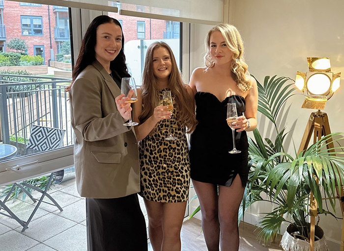 Three women dressed up carrying wine glasses surrounded by greenery