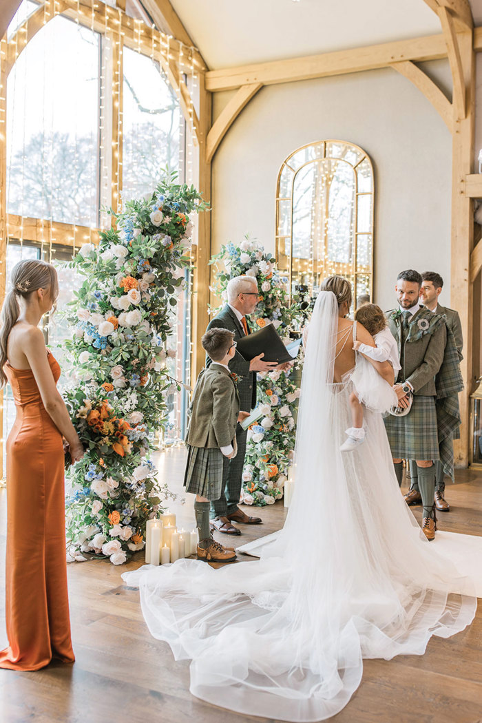 large floral half arch at the front of wedding ceremony with bride in long train and veil, holding young girl in white outfit, and groom and groomsmen in green and blue tartan outfits 