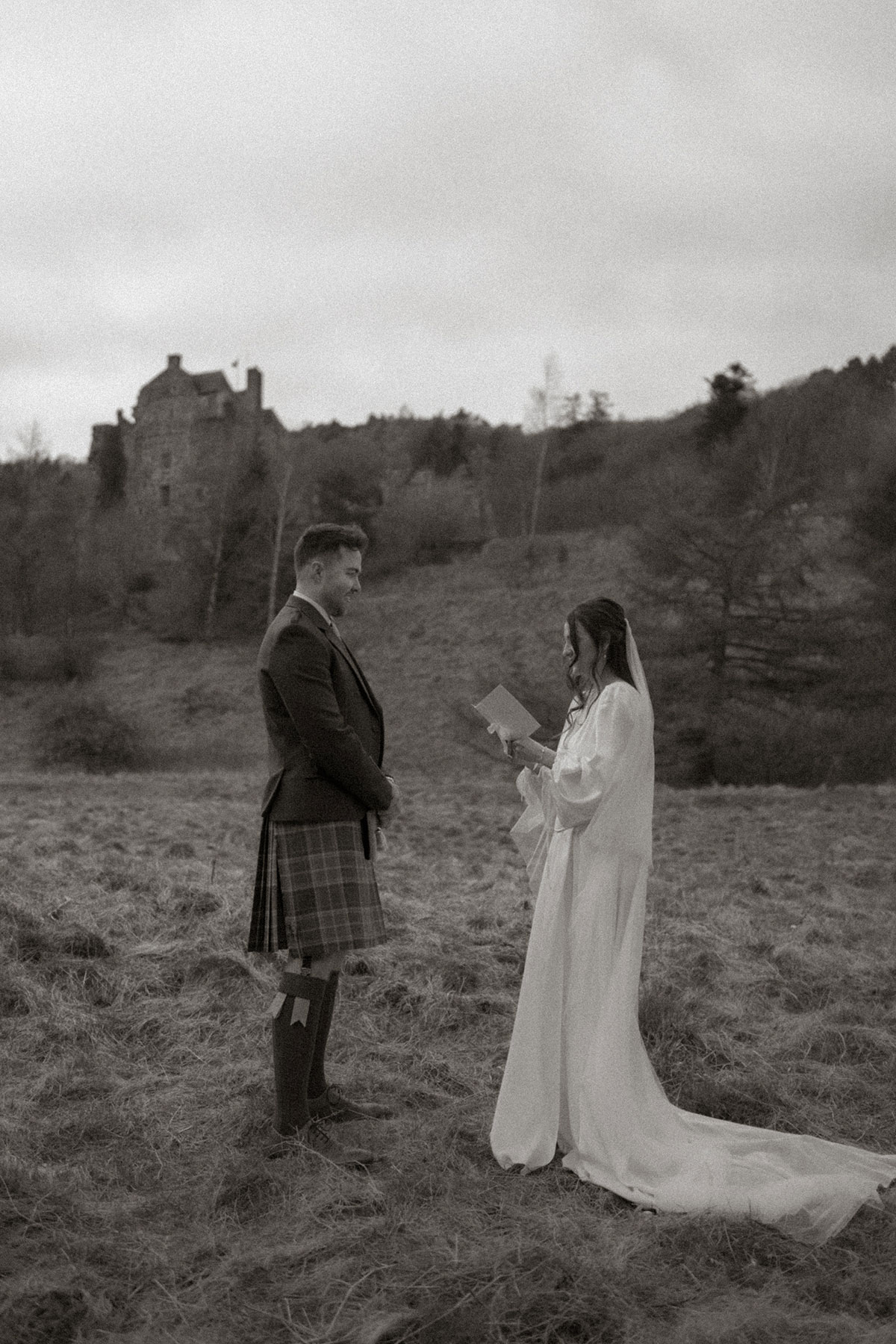 black and white image of bride in trailing satin dress with matching veil reading from a booklet, standing facing a groom in kilt outfit, stood outdoors on the grass with a small blurred castle in the background