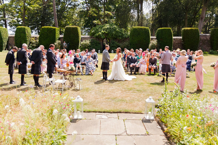 an outdoor wedding ceremony with guests sitting on chairs on the lawn while the groomsmen, bridesmaids, bride and groom are standing 