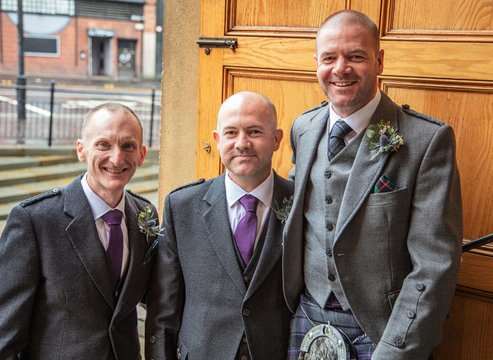 Three men in grey suits smile facing the camera