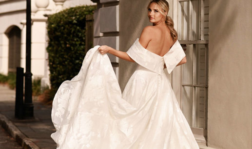 bride wearing large dramatic off-the-shoulder wedding dress looks back over her shoulder while holding up one corner of her large ballgown skirt