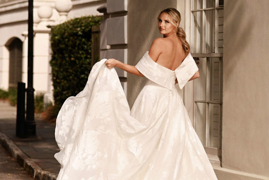 bride wearing large dramatic off-the-shoulder wedding dress looks back over her shoulder while holding up one corner of her large ballgown skirt