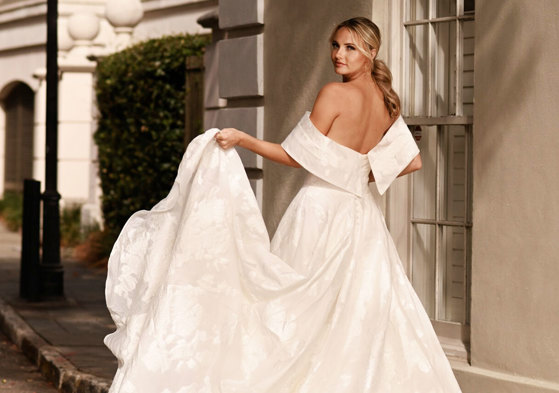 bride wearing large dramatic off-the-shoulder wedding dress looks back over her shoulder while holding up one corner of her large ballgown skirt