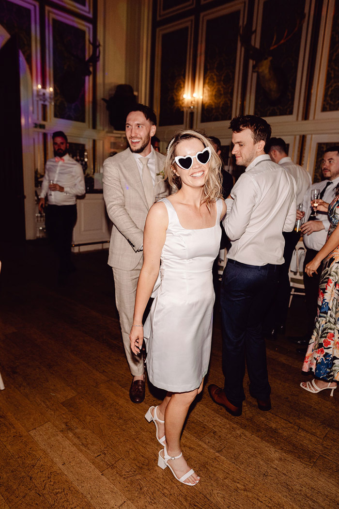 bride leads groom through evening reception with guests while wearing heart shaped glasses