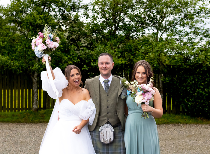 bride standing in white dress holding flowers above head, groom in kilt and young girl in teal dress