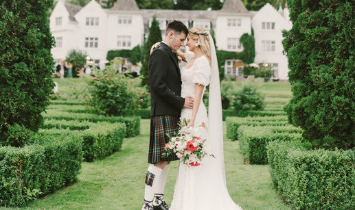 A bride and groom stand outside facing each other in a garden with small hedges and fir trees in front of their venue