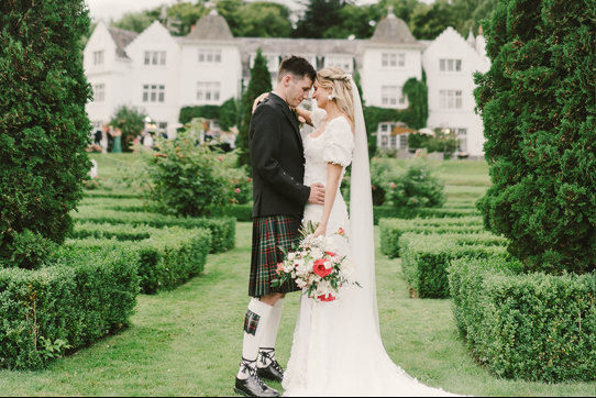 A bride and groom stand outside facing each other in a garden with small hedges and fir trees in front of their venue