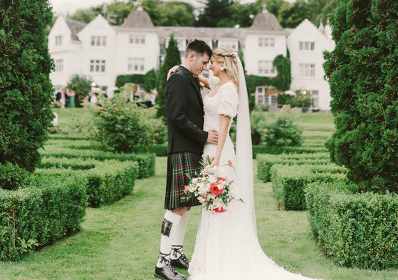 A bride and groom stand outside facing each other in a garden with small hedges and fir trees in front of their venue