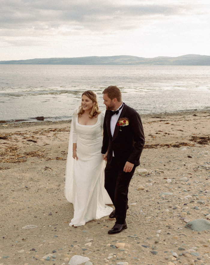 a bride and groom walking hand in hand along a stone pebble beach