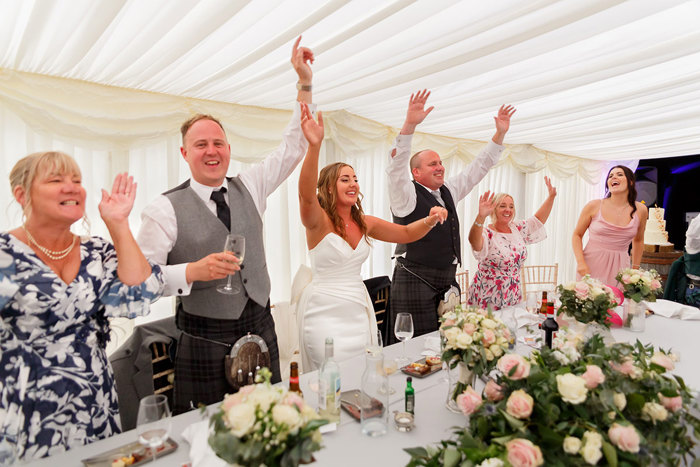 Top table at a wedding with six people cheering with their hands in the air and a white curtain backdrop