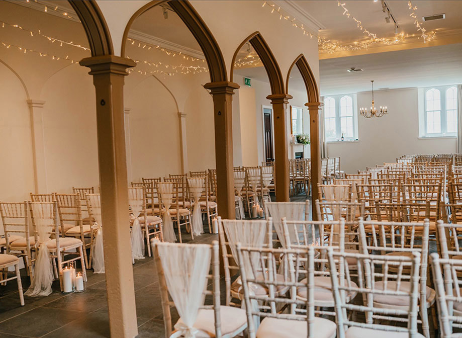 A room with lots of wooden arch ways set up for a wedding ceremony with white chiavari chairs draped in white fabric 