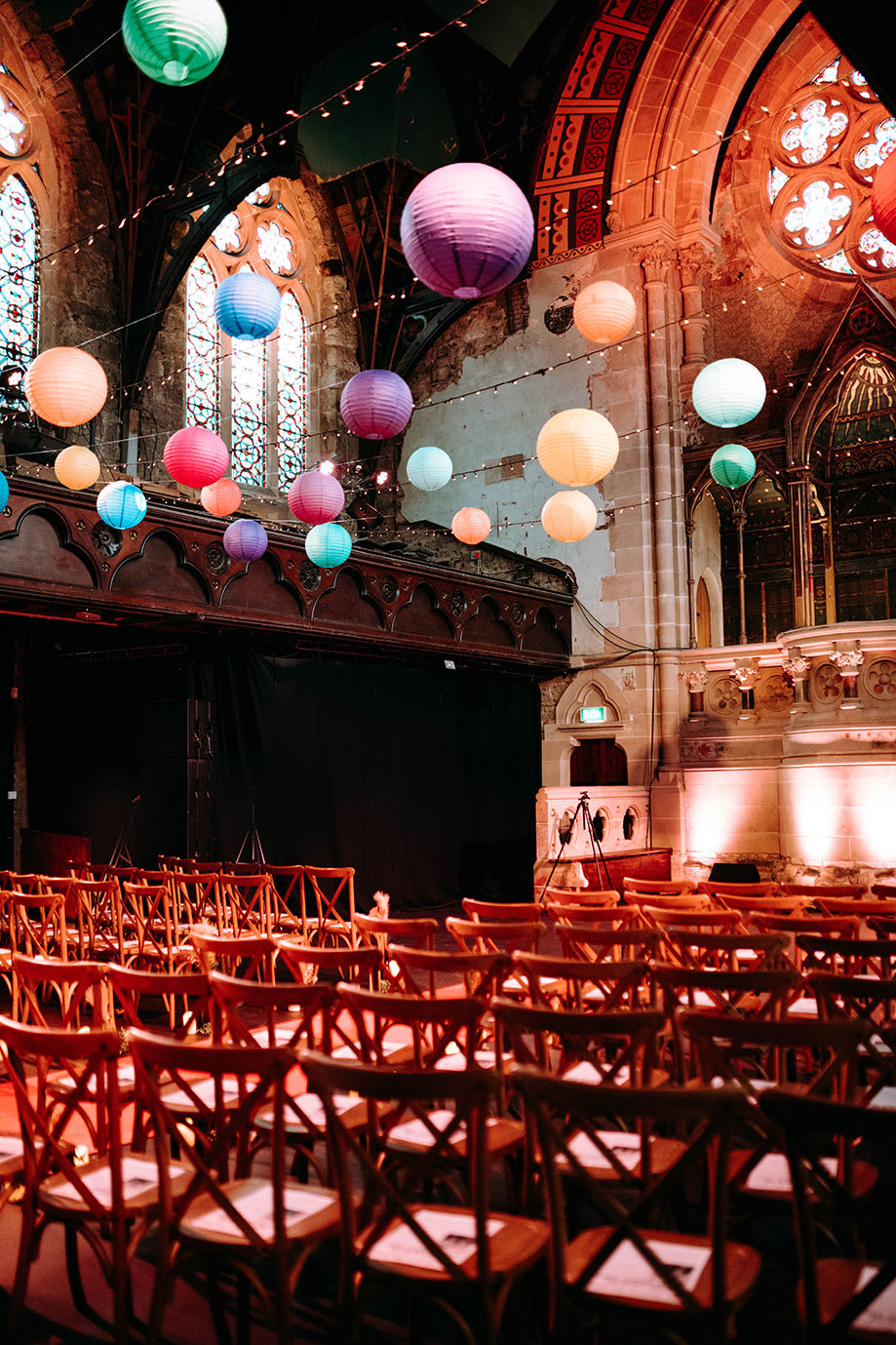 rows of wooden cross back chairs set out for a wedding ceremony in a church style building with rows of colourful hanging lanterns on left. A bride and groom walking along a road outside a tall ornate church building on right