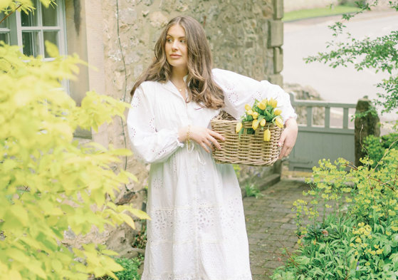 a brunette woman in a long white dress stands outside a stone house holding a basket of yellow tulips