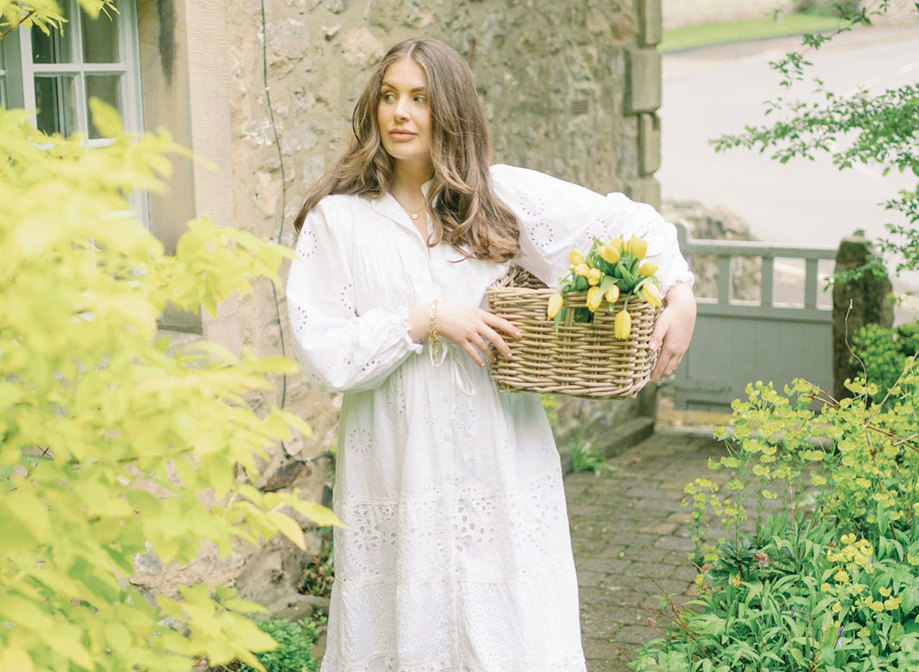 a brunette woman in a long white dress stands outside a stone house holding a basket of yellow tulips