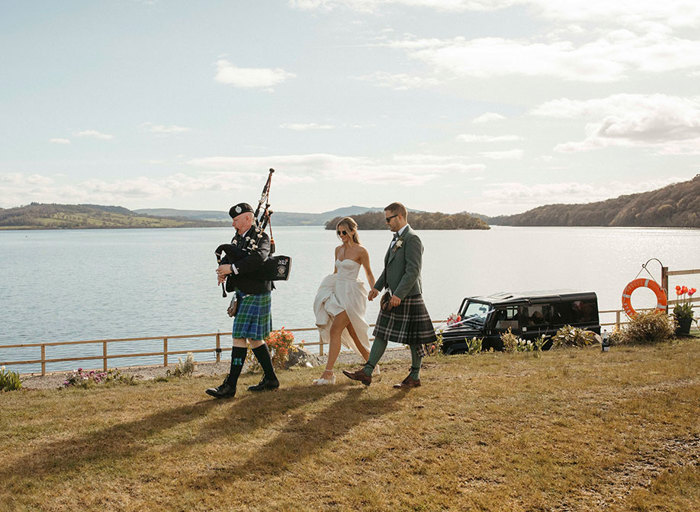 a bride and groom walking along behind a man playing the bagpipes in front of Loch Lomond