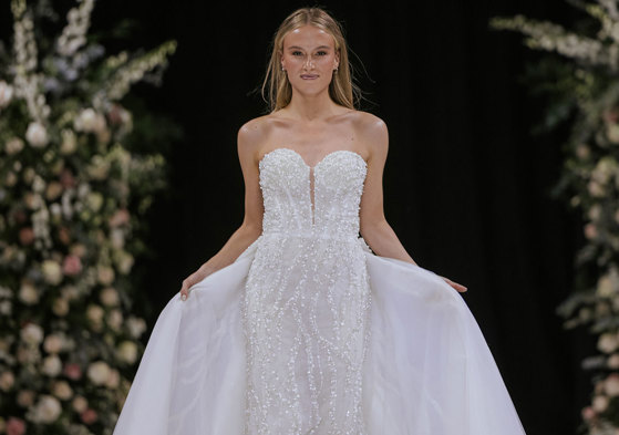 a model wearing a white strapless wedding dress on a catwalk. There are two tall flower displays in the background