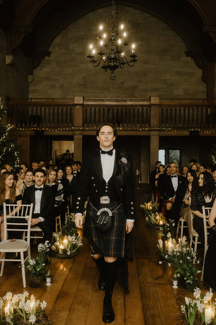 a man in a kilt walking down an aisle as rows of seated wedding guests look on.