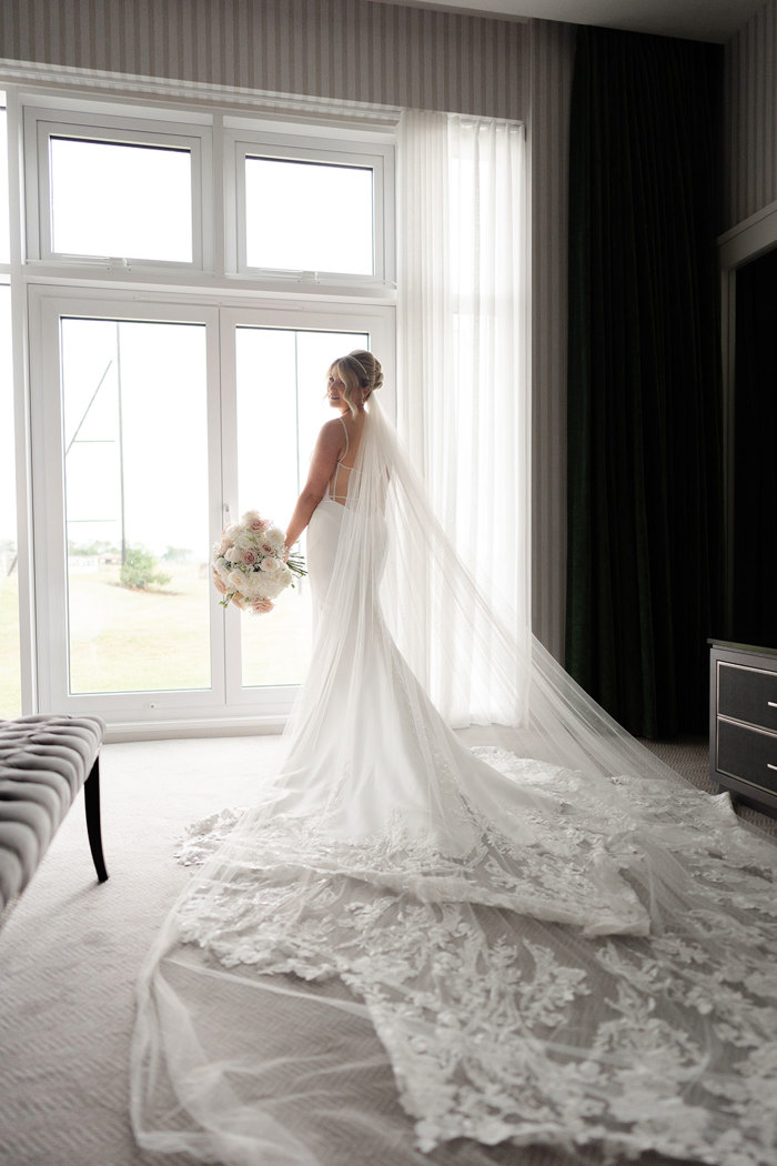 a bride by a window in a dress with long lace train and veil.